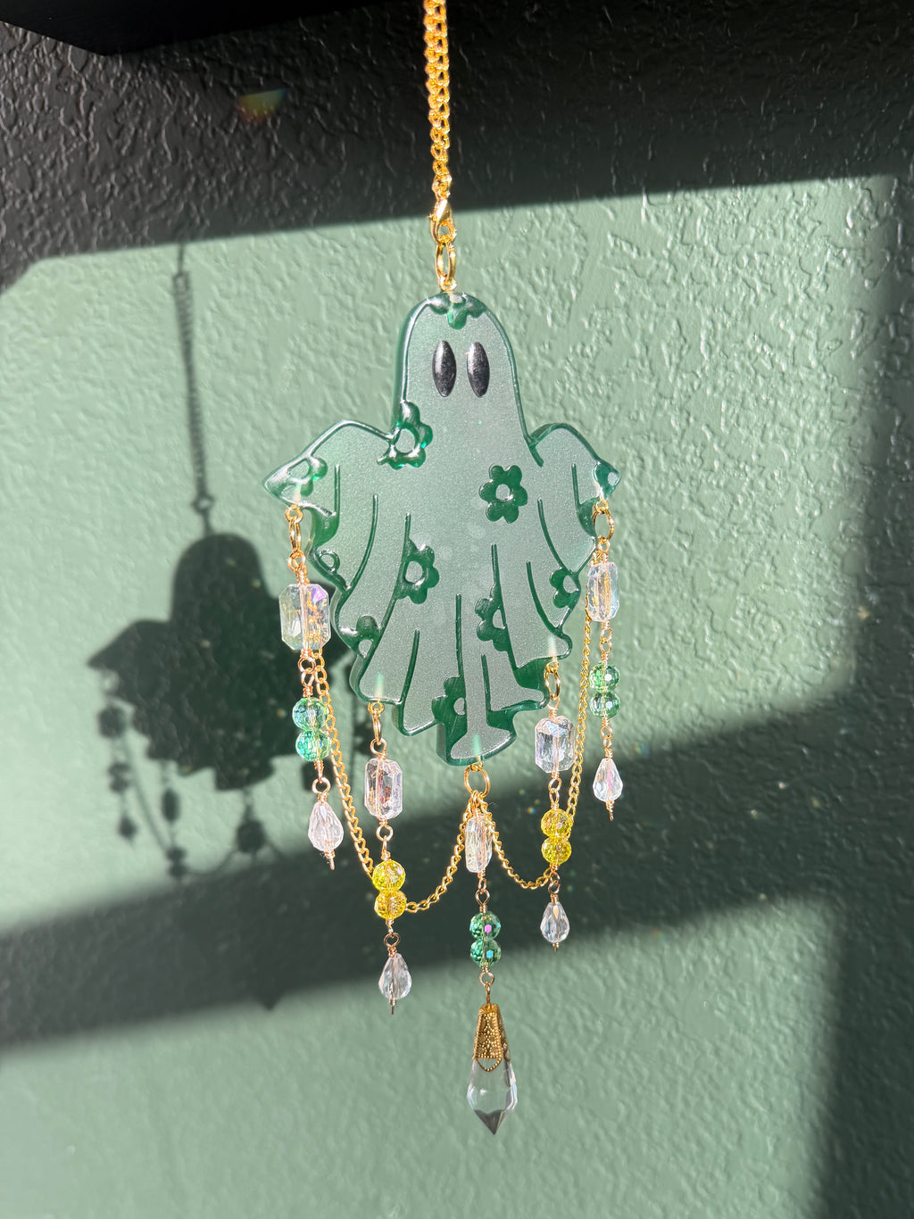 A decorative hanging ornament shaped like a ghost, made from translucent pale green material with green floral patterns, black eyes, and dangling gold chains with clear and colored beads, shown against a painted green wall with strong shadows.