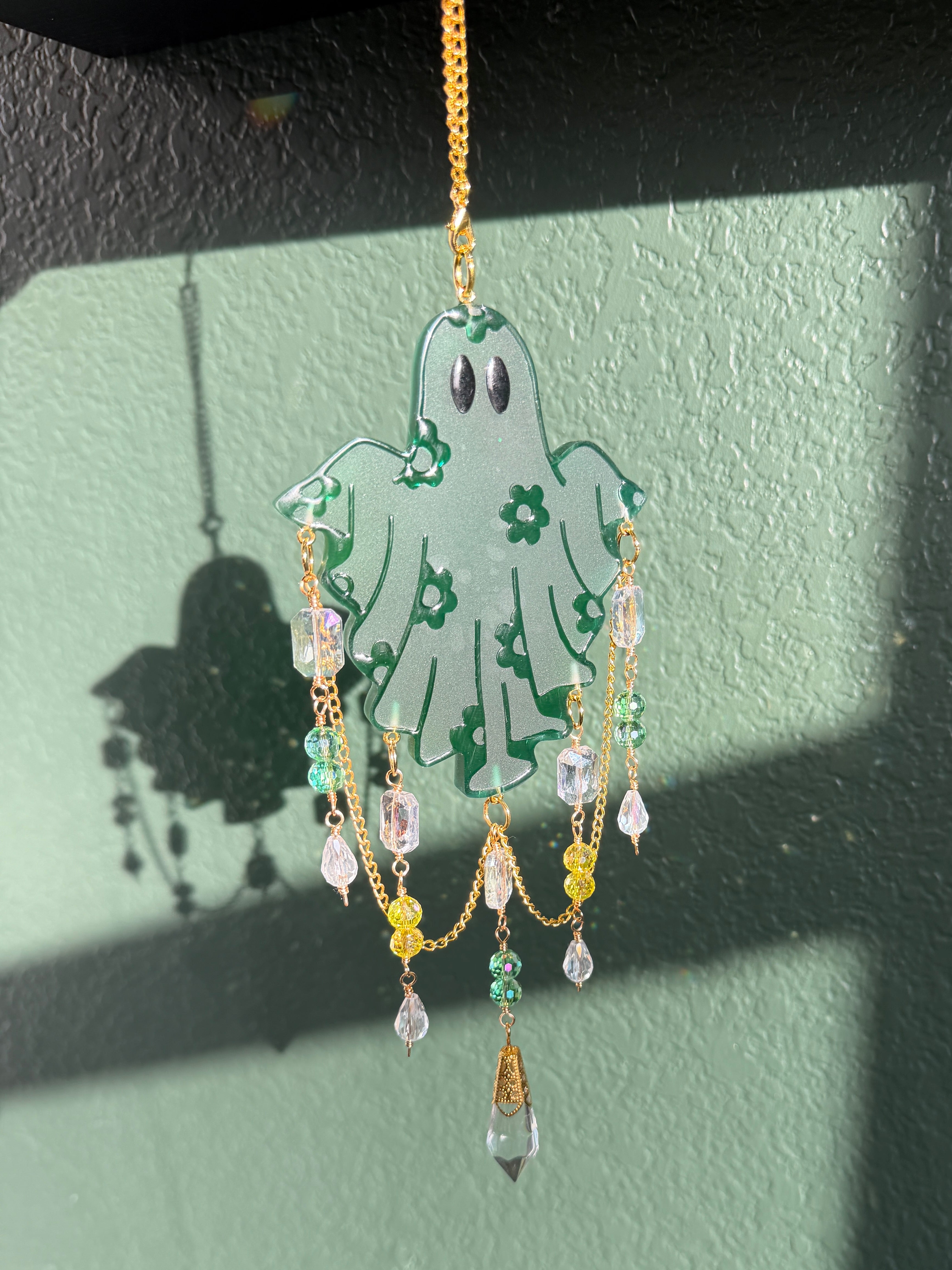 A decorative hanging ornament shaped like a ghost, made from translucent pale green material with green floral patterns, black eyes, and dangling gold chains with clear and colored beads, shown against a painted green wall with strong shadows.