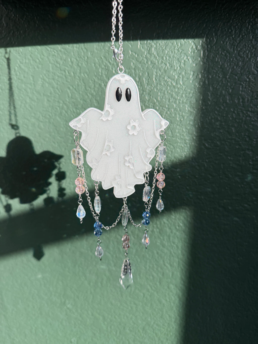 A white ghost-shaped pendant  with black eyes, decorated with small white floral details and dangling clear and pastel beads on a silver chain, shown against a textured green wall.