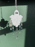 A white ghost-shaped pendant  with black eyes, decorated with small white floral details and dangling clear and pastel beads on a silver chain, shown against a textured green wall.