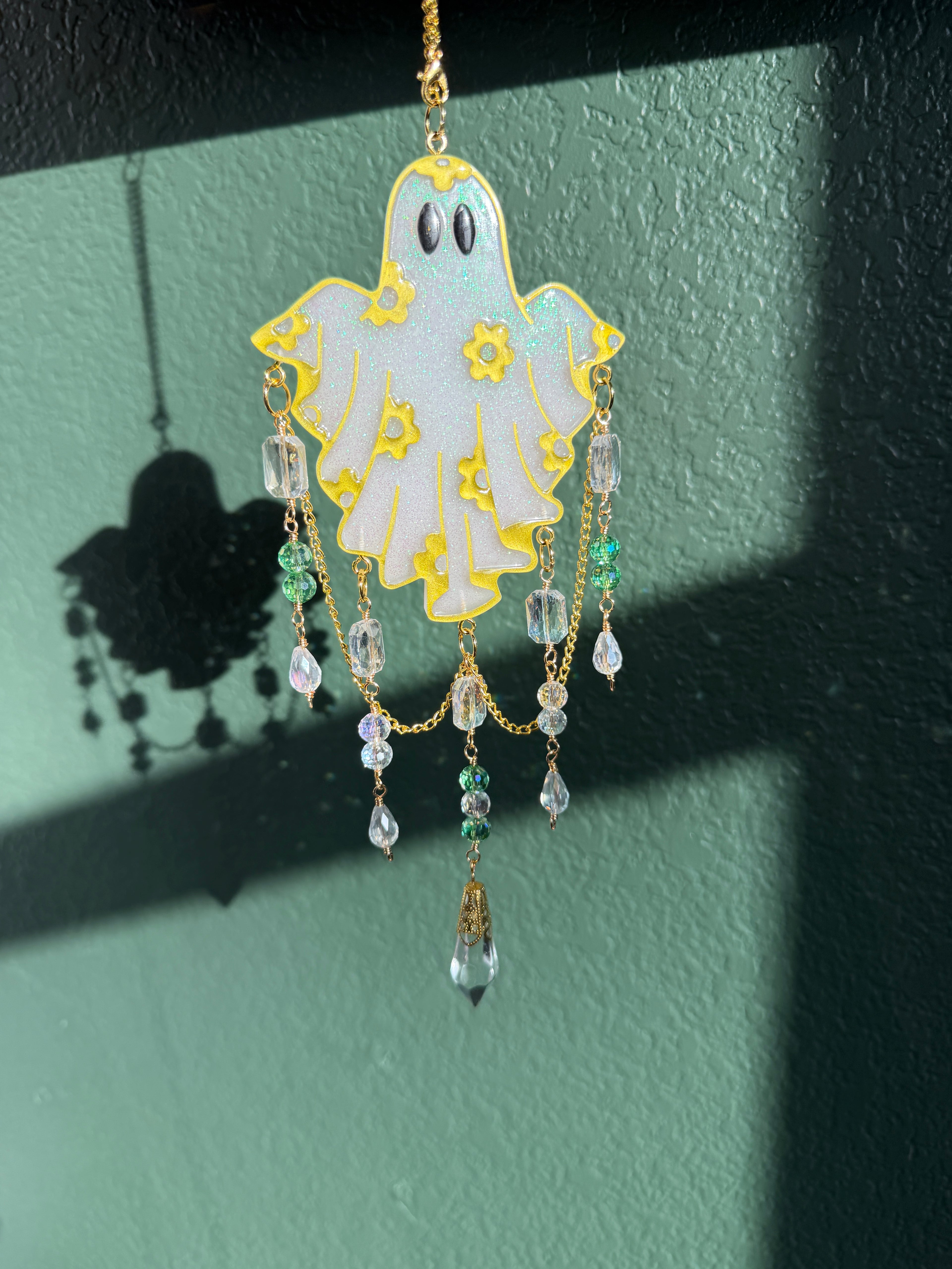A decorative ghost-shaped wall hanging ornament with gold trim, a white body, black eyes, and dangling clear and green bead chains, shown against a dark green wall with strong sunlight and shadows.