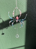 A handmade beaded spider ornament hanging from a chain against a textured green wall, featuring pink, light blue, and white beads with a clear faceted crystal pendant.