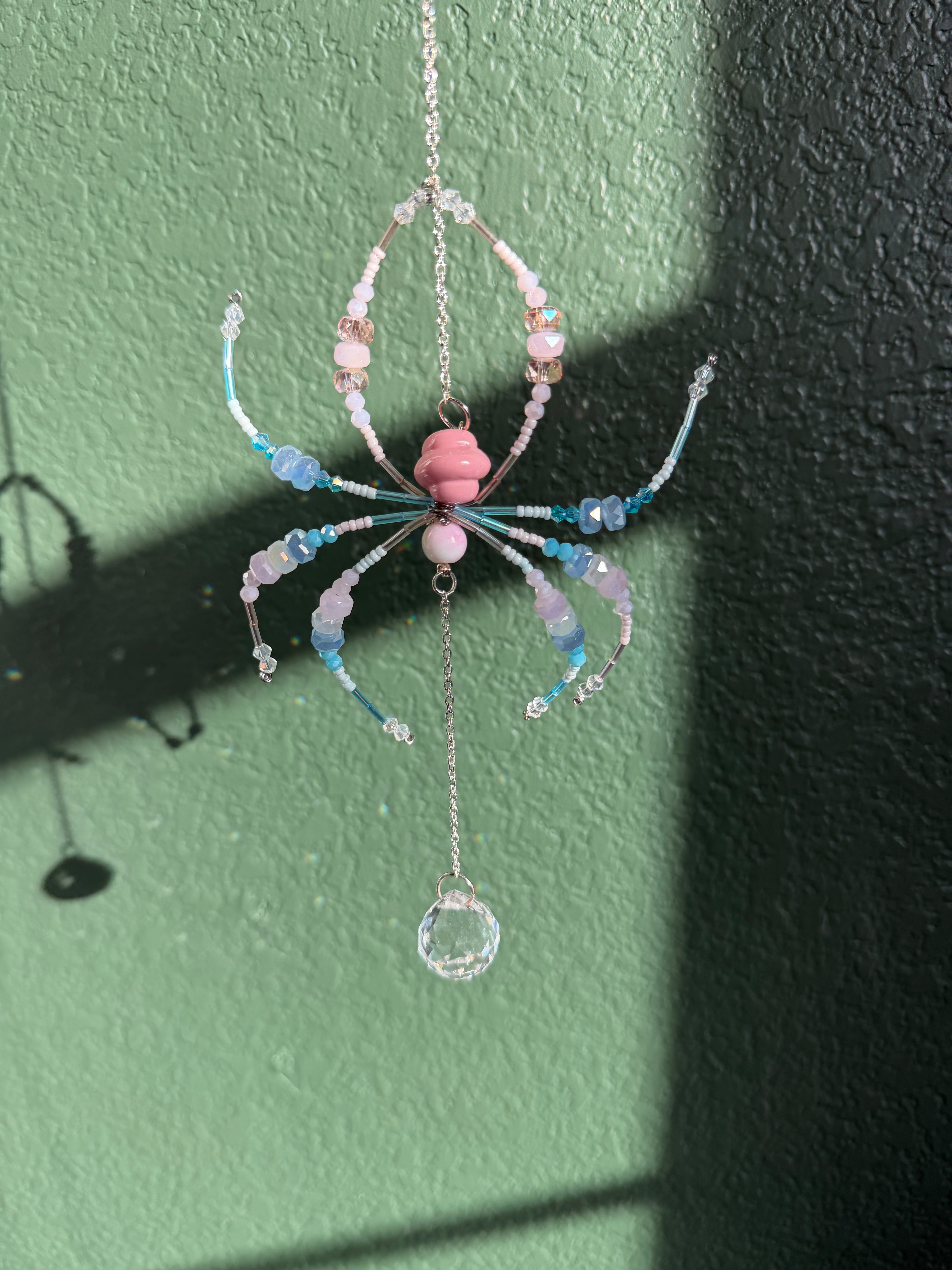 A handmade beaded spider ornament hanging from a chain against a textured green wall, featuring pink, light blue, and white beads with a clear faceted crystal pendant.