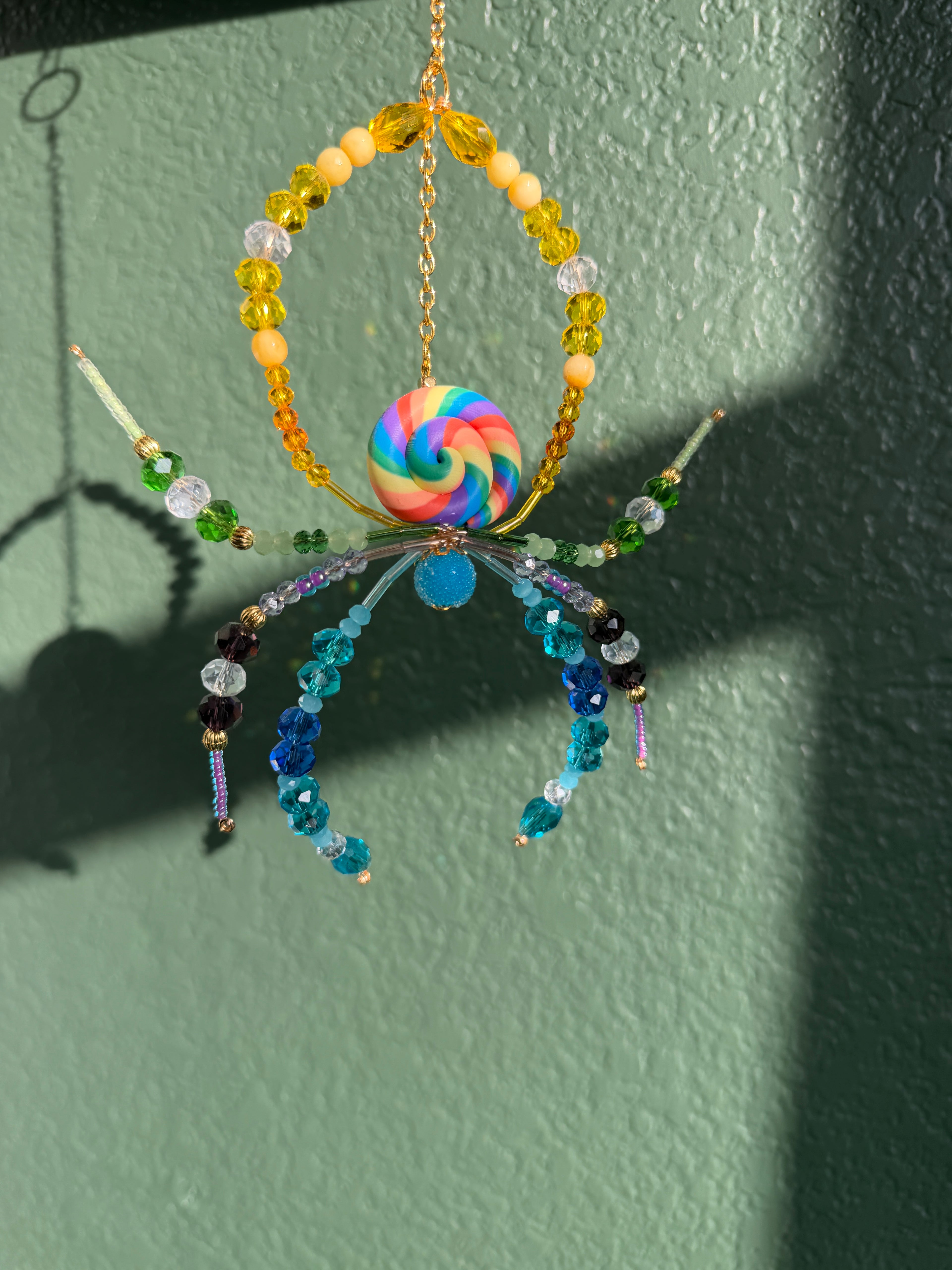 A colorful handmade beaded hanging ornament shaped like a spider or crab, featuring a rainbow swirl center bead, yellow and blue beads, and translucent beads, hanging against a textured green wall with strong sunlight and shadows.