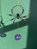 A hanging spider ornament made with dark faceted beads and a gold chain, with a purple circular tag below that reads 'Feral'. The ornament is photographed against a textured green wall with strong sunlight and shadows.