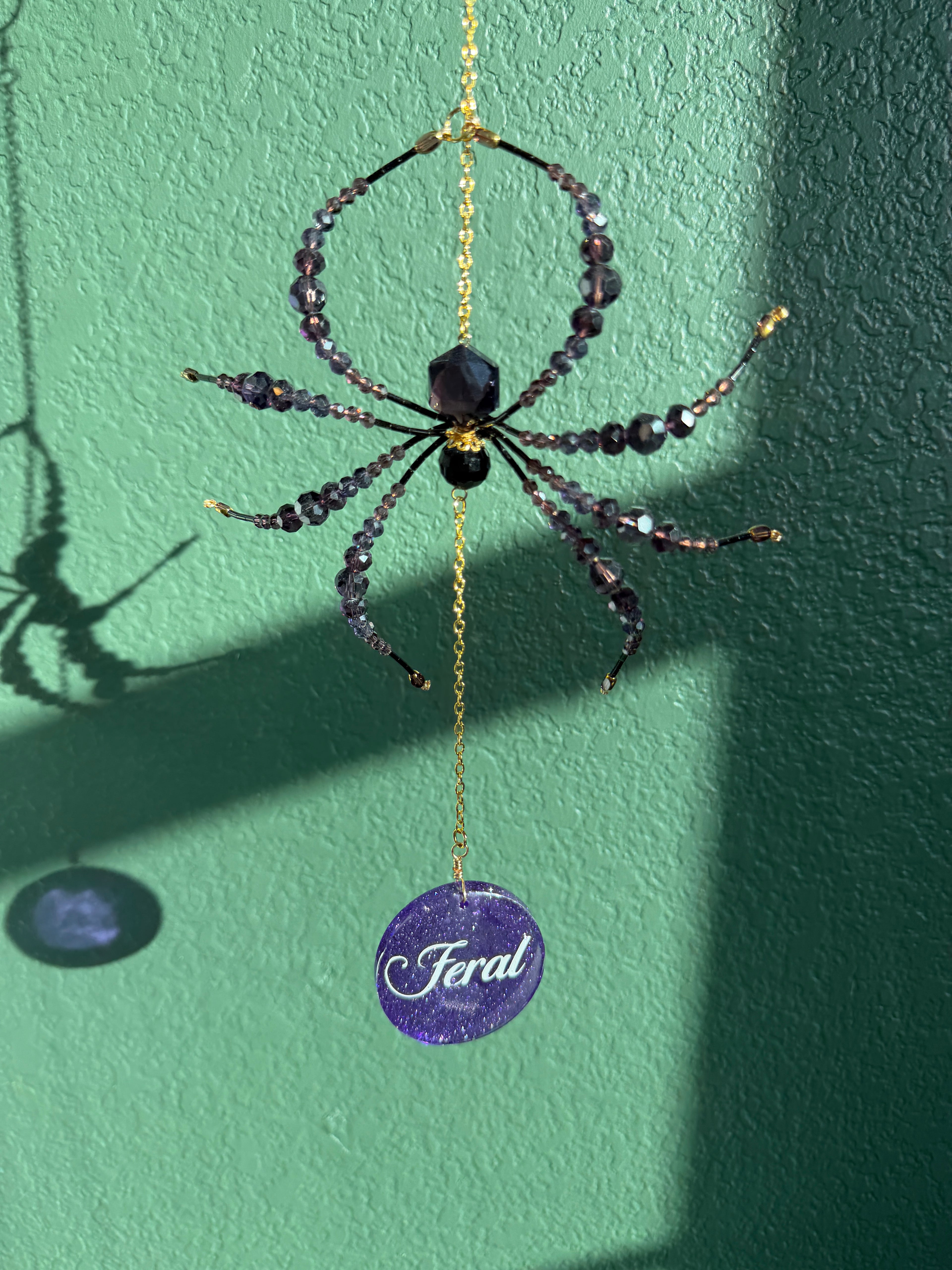 A hanging spider ornament made with dark faceted beads and a gold chain, with a purple circular tag below that reads 'Feral'. The ornament is photographed against a textured green wall with strong sunlight and shadows.