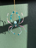 A handmade beaded spider ornament hanging from a gold chain against a textured green wall. The spider features light blue and pink beads, a light blue center bead, and gold-toned metal details.