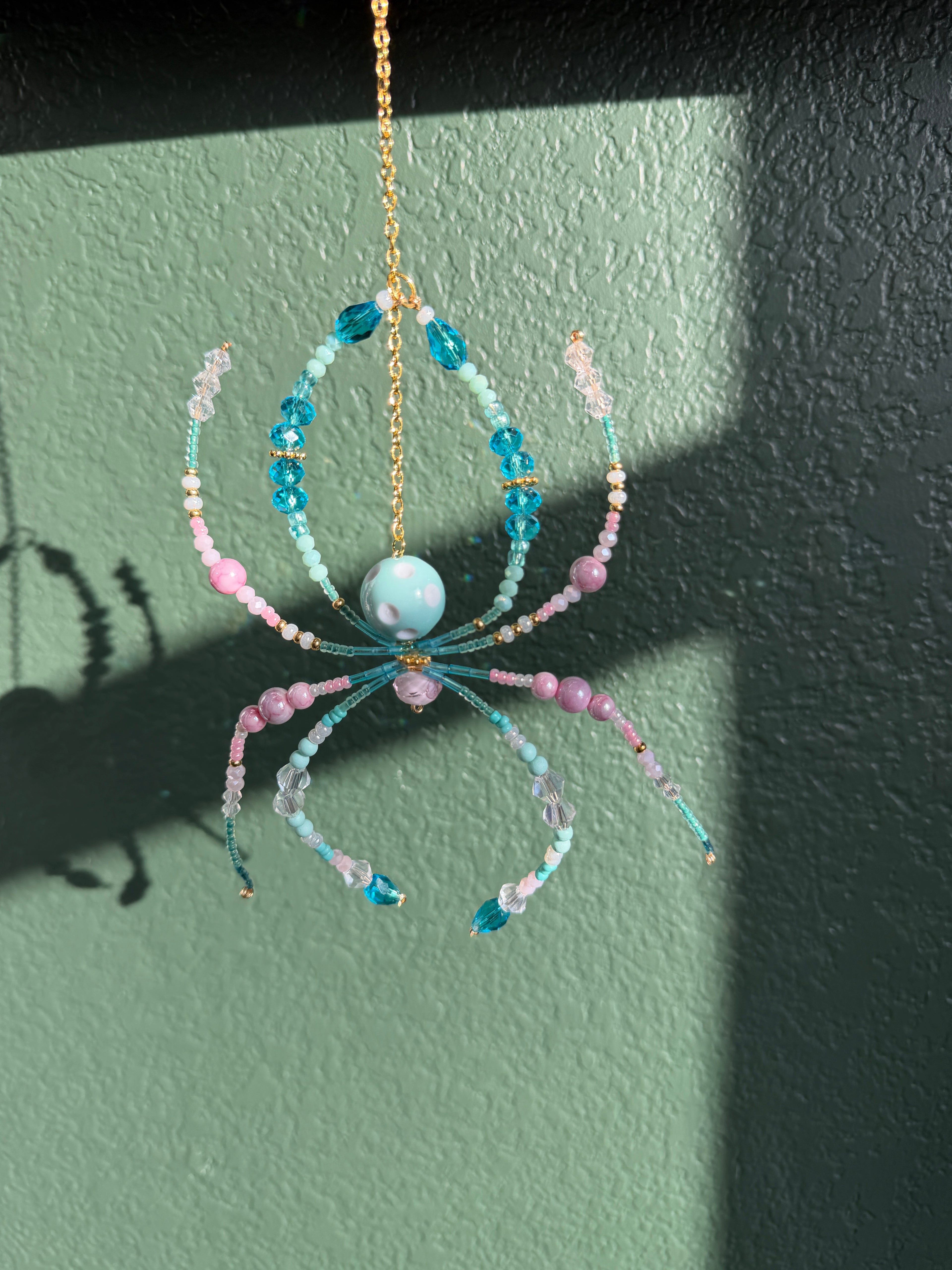 A handmade beaded spider ornament hanging from a gold chain against a textured green wall. The spider features light blue and pink beads, a light blue center bead, and gold-toned metal details.