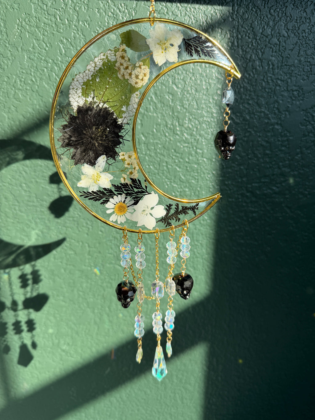 A gold crescent moon wall hanging ornament decorated with pressed flowers, green leaves, and hanging beaded charms, displayed against a textured green wall with natural sunlight and shadows.