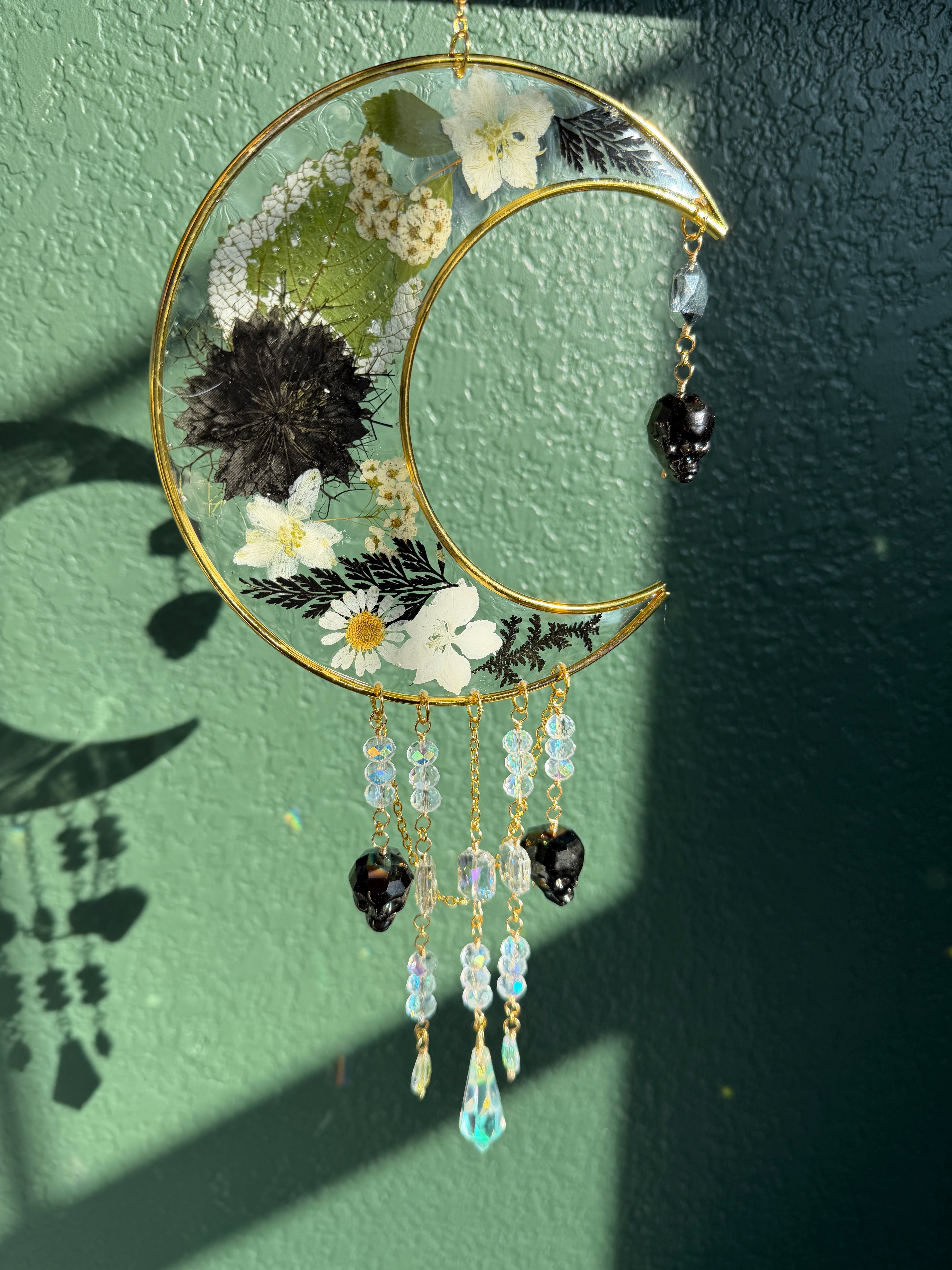 A gold crescent moon wall hanging ornament decorated with pressed flowers, green leaves, and hanging beaded charms, displayed against a textured green wall with natural sunlight and shadows.