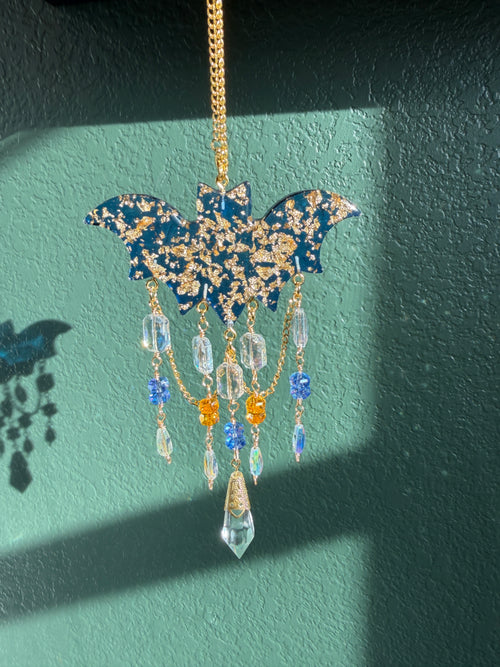 A gold chain necklace featuring a bat-shaped pendant decorated with blue and gold glitter, adorned with dangling clear faceted beads and small blue and orange charms, set against a textured teal wall with a shadow.
