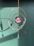 A handmade circular hoop wall hanging ornament with rose gold metal wire, clear faceted beads, and a dangling iridescent crystal. The center features a circular sticker that reads 'Be KIND or a BITCH' in pink and white text.