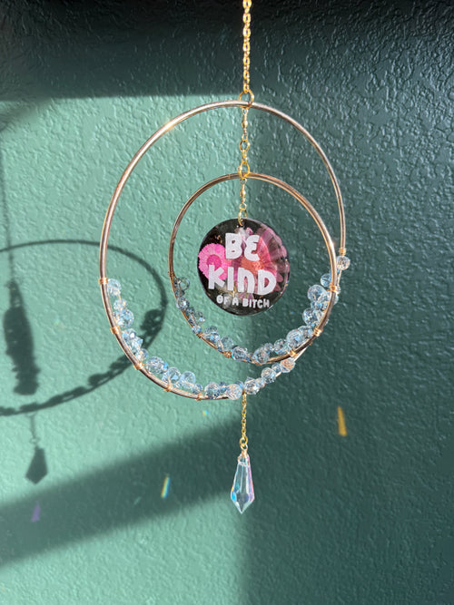 A handmade circular hoop wall hanging ornament with rose gold metal wire, clear faceted beads, and a dangling iridescent crystal. The center features a circular sticker that reads 'Be KIND or a BITCH' in pink and white text.